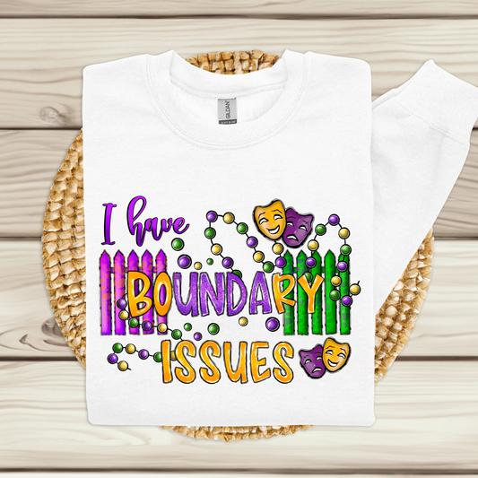 A white hooded sweatshirt with a colorful design featuring the text 'I have BOUNDARY ISSUES' and related to Mardi Gras theme, displayed on a wooden background.
