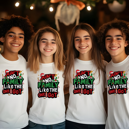 Four children wearing white t-shirts with a Christmas-themed design, standing in front of a decorated Christmas tree.