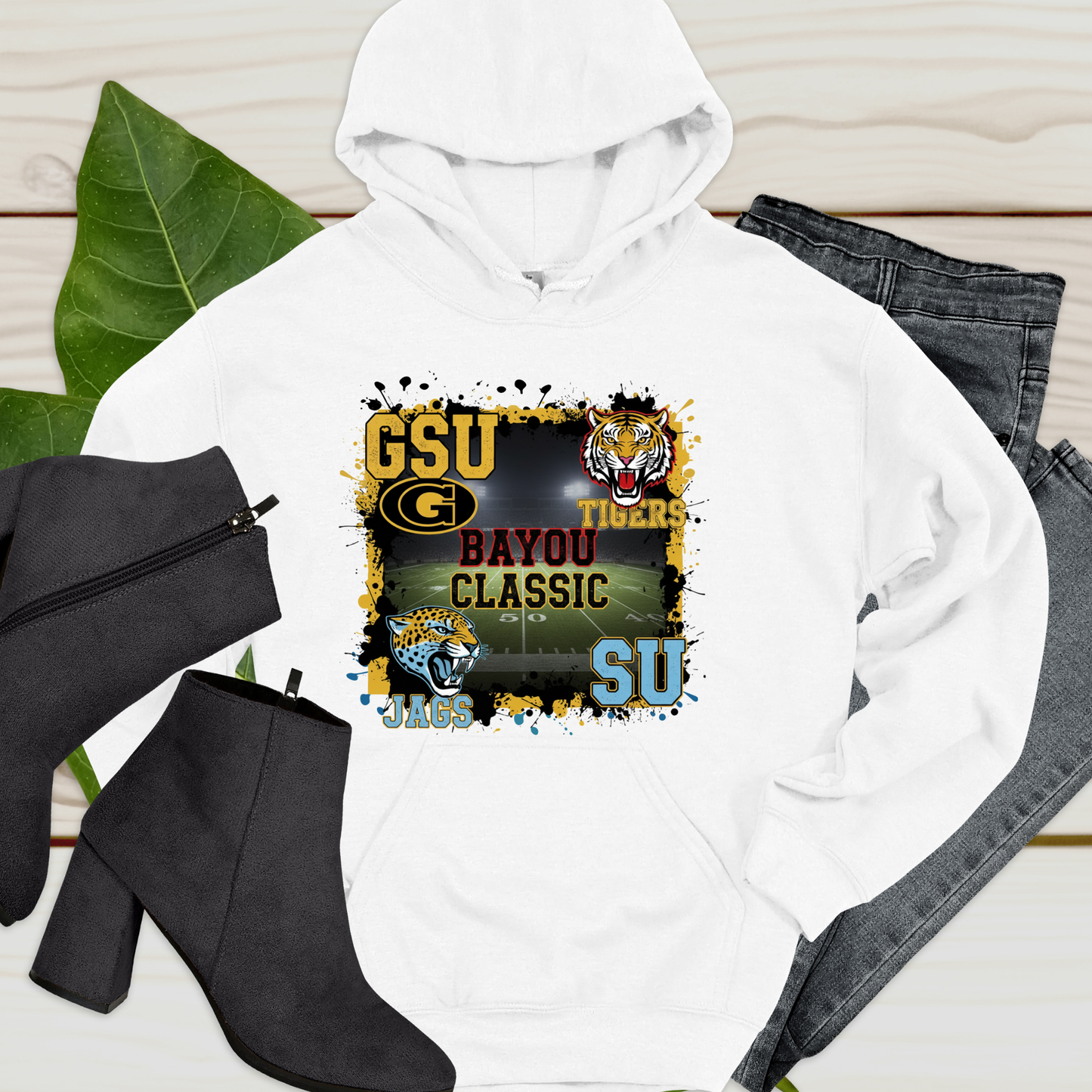 White hoodie with sports-themed design, black boots, and jeans on a wooden surface. Bayou Classic Football Hoodie - Grambling State GSU Tigers vs. Southern University SU Jags Hoodie