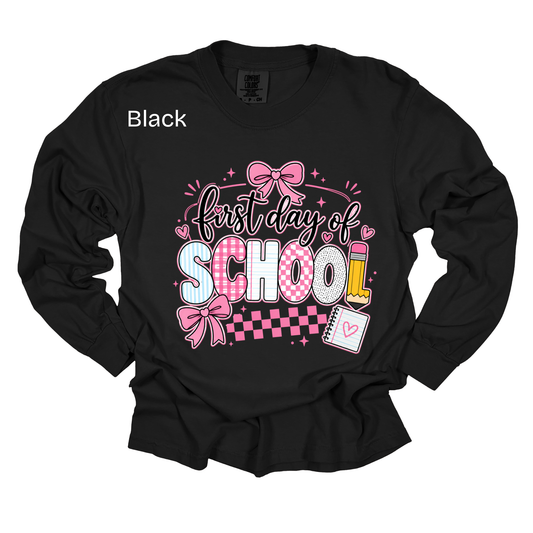 A black long-sleeve t-shirt with a 'First day of SCHOOL' graphic on the front, featuring pink bows, checkered patterns, and school-themed illustrations. The word 'Black' is printed in white on the upper left chest.