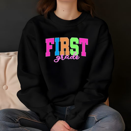 A person wearing a black crewneck sweatshirt with a colorful graphic that reads 'FIRST grade' in bright pink, blue, orange, and green block letters, paired with blue jeans, sitting against a dark background.