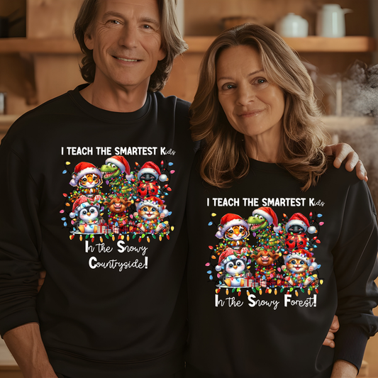 A man and a woman wearing matching black long-sleeve crewneck sweatshirts with a colorful Christmas cartoon graphic and the text 'I TEACH THE SMARTEST Kids In the Snowy Countryside! / In the Snowy Forest!'. The man wears 'Countryside' and the woman wears 'Forest' in the graphic text.