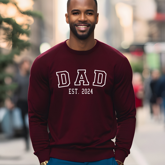 Man wearing a maroon sweatshirt with "DAD" in bold letters and "EST. 2024" below, featuring a heart graphic with kid names personalized on the sleeve. New Dad sweater.