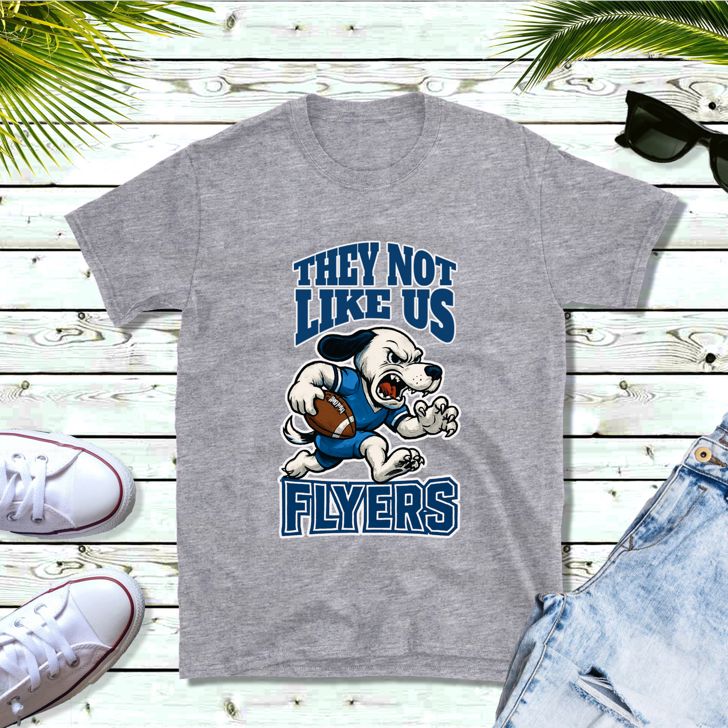 Gray t-shirt with cartoon character and text on a wooden surface with shoes and shorts. Flyers Mascot T-Shirt: "They Not Like Us" Flyers Game Day Graphic Tee