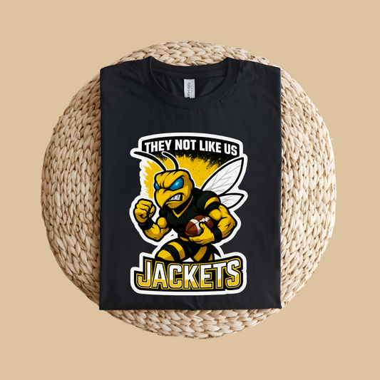 Black t-shirt with a yellow and black graphic of a bee holding a football, text 'THEY NOT LIKE US JACKETS', on a woven mat. Jackets Mascot Shirt - Bold Yellow Jacket They Not Like Us Spirit Tee