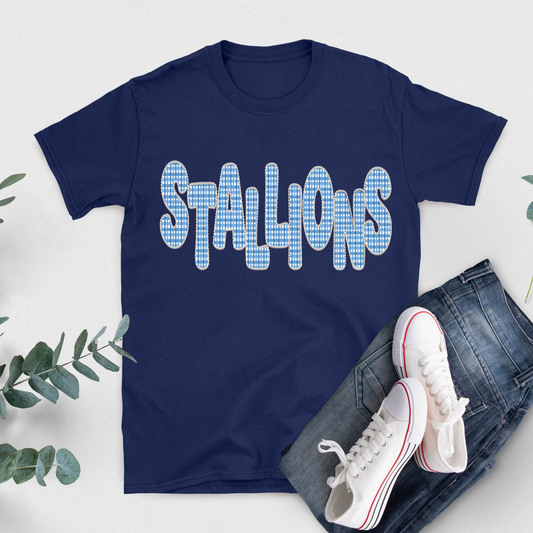 A navy blue short-sleeve t-shirt with the word 'STALLIONS' printed across the chest in a light blue and white gingham pattern. The shirt is styled in a flat lay with blue jeans, white sneakers, and green eucalyptus leaves on a light gray background.