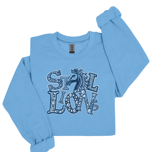 A light blue long-sleeve crewneck sweatshirt featuring a graphic print of the word 'STILL LOVE' in decorative letters with a horse silhouette in the center, displayed on a clean white background.