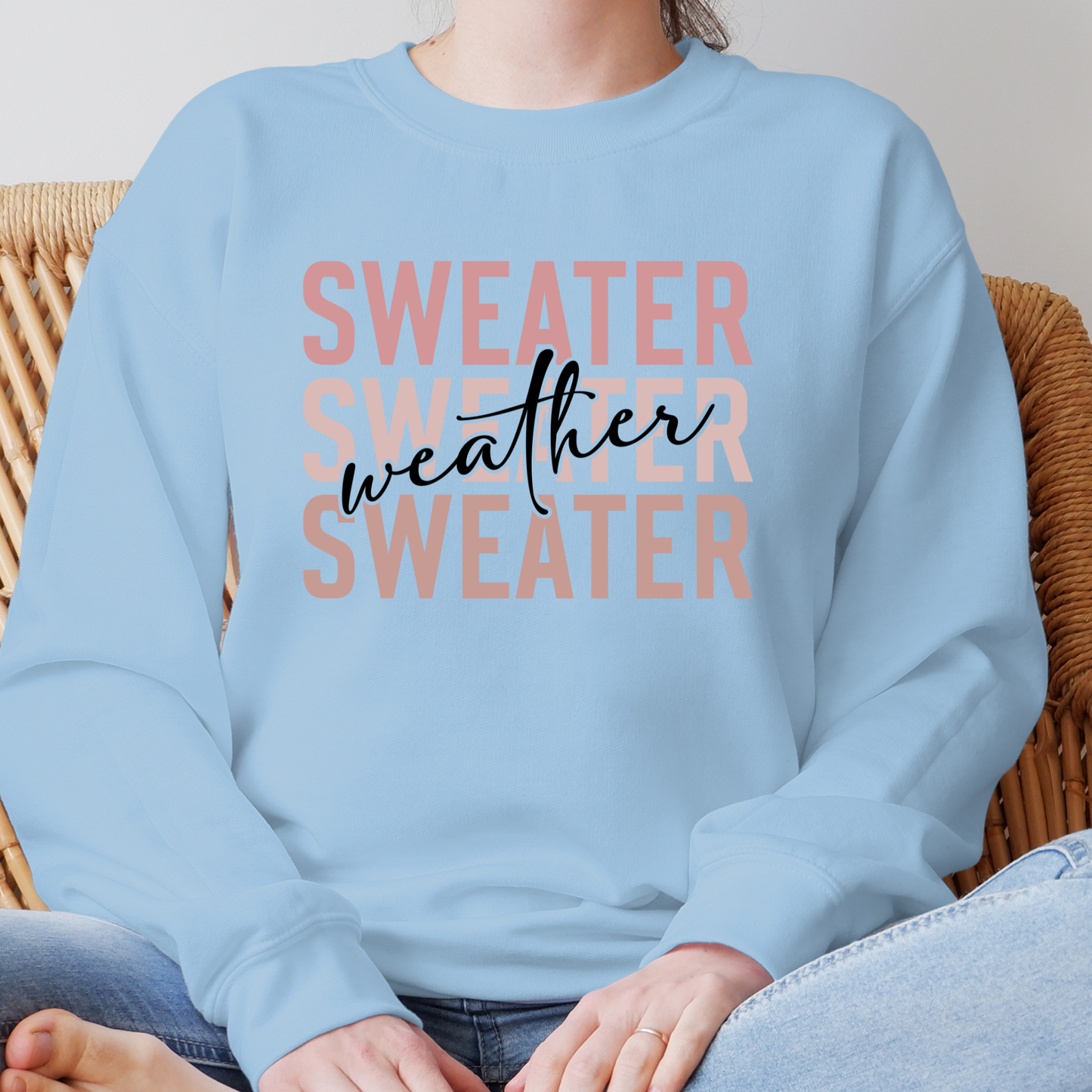 Light blue sweatshirt with 'SWEATER WEATHER SWEATER' text worn by a person sitting on a wicker chair. Sweater Weather - Cute and Chic Minimalist Fall Crewneck Cozy Sweater