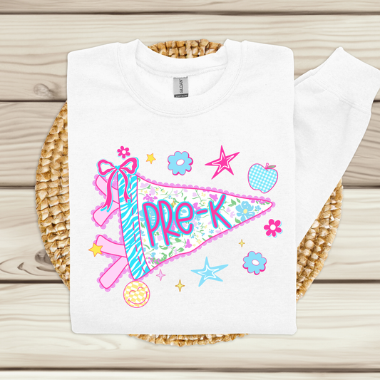 A white long-sleeve sweatshirt laid flat on a woven round mat over a light wood plank surface. The front has a colorful graphic of a pink and blue striped pennant flag with the word 'Pre-K' in purple, surrounded by flowers, stars, and an apple.