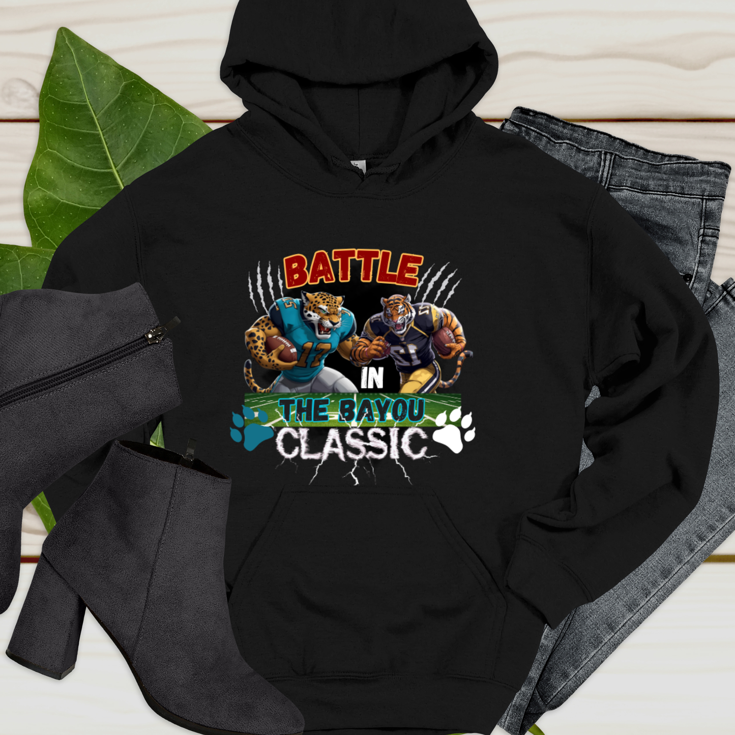 Black hoodie with 'Battle in the Bayou Classic' graphic on a wooden surface with accessories. Bayou Classic Hoodie: Battle in the Bayou Classic Game Day Unisex Hoodie