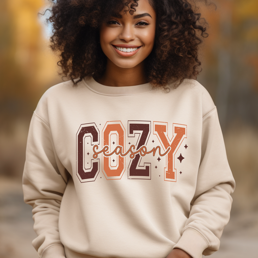 Person wearing a beige sweatshirt with 'COZY season' text in an autumn setting Cozy Season Sweatshirt: Fall Vibe Retro Block Letter Crewneck Sweater