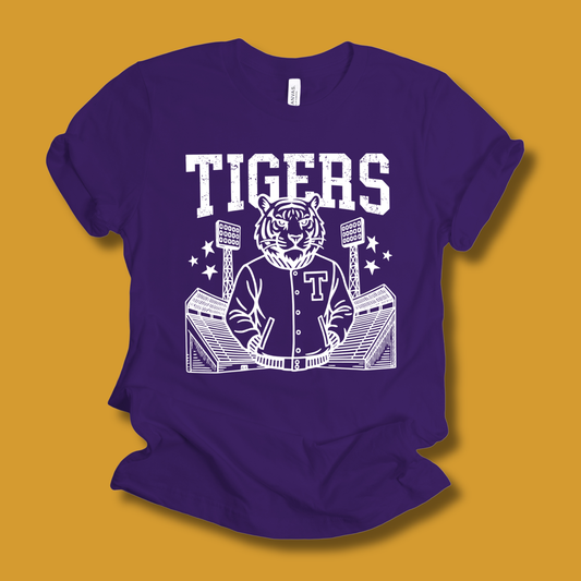 A purple short-sleeve t-shirt with a white graphic print of a tiger mascot in a varsity jacket, stadium lights, and the word 'TIGERS' on the front, displayed against a solid mustard yellow background.