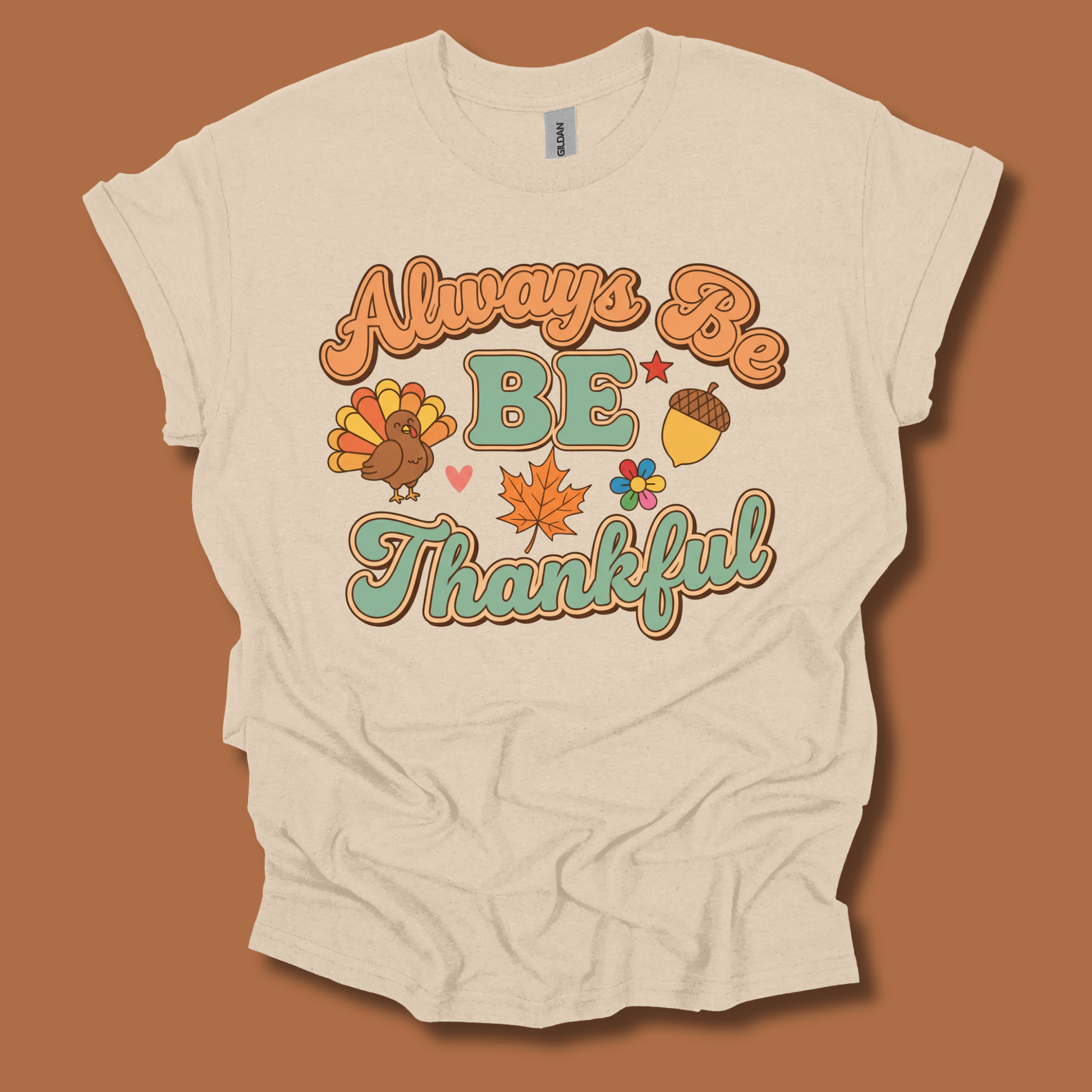 A cream-colored short-sleeve t-shirt with a retro-style autumn graphic that reads 'Always Be BE Thankful' in orange and teal lettering, accompanied by a turkey, acorn, maple leaf, and small flowers. The shirt is displayed flat against a solid terracotta brown background.