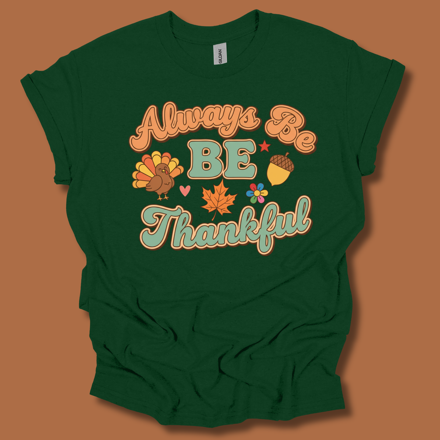 A dark green short-sleeve t-shirt with a retro-style Thanksgiving graphic that reads 'Always Be BE Thankful' in orange and teal lettering, accompanied by a turkey, acorn, maple leaf, and flowers, displayed against a solid brown background.