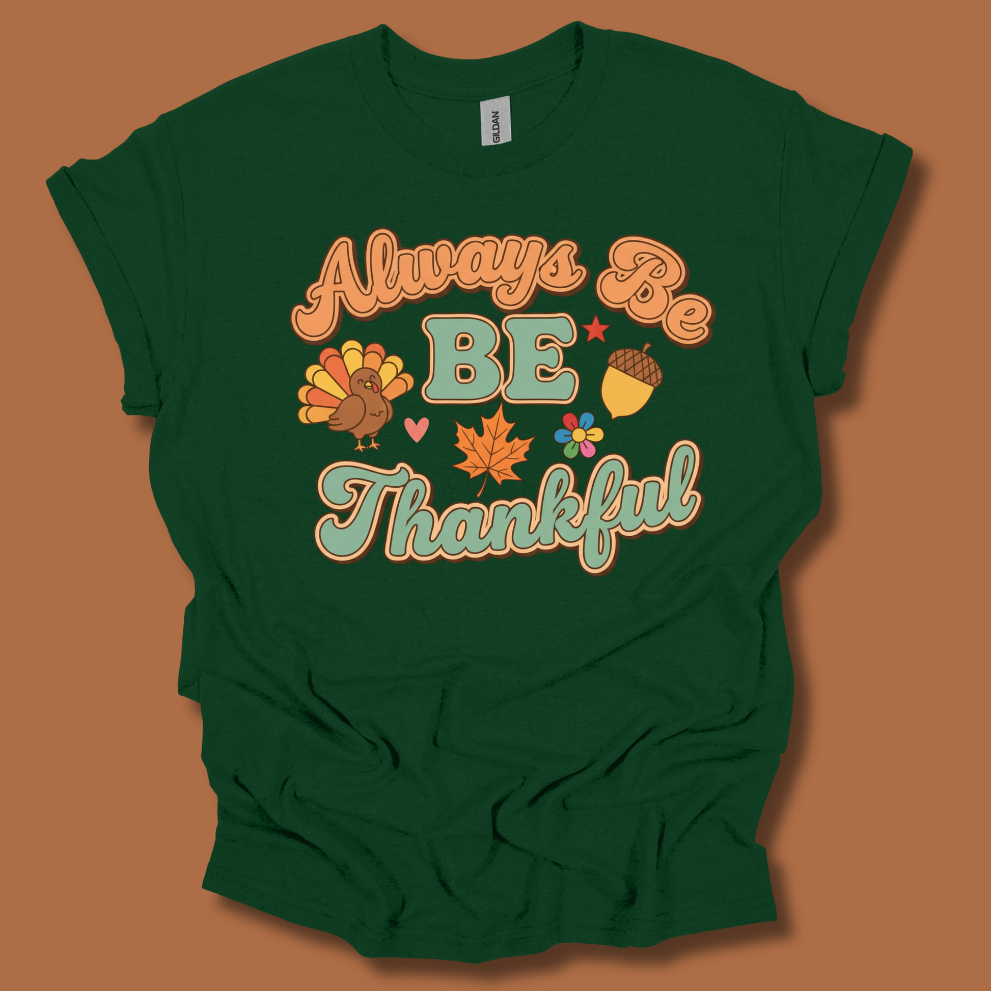 A dark green short-sleeve t-shirt with a retro-style Thanksgiving graphic that reads 'Always Be BE Thankful' in orange and teal lettering, accompanied by a turkey, acorn, maple leaf, and flowers, displayed against a solid brown background.