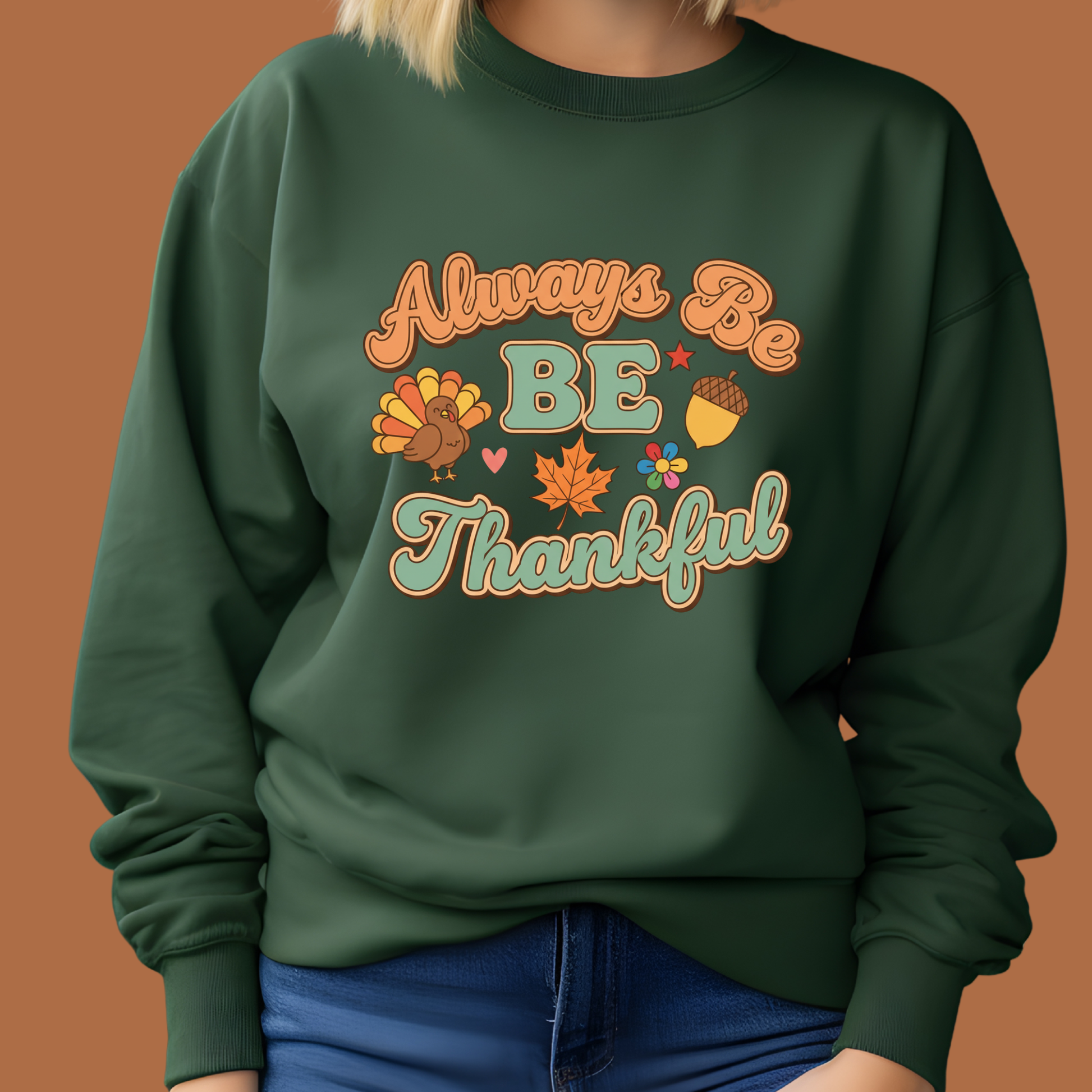 A person wearing a dark green crewneck sweatshirt with a colorful 'Always Be Thankful' Thanksgiving graphic on the front, paired with blue jeans against a solid brown background.