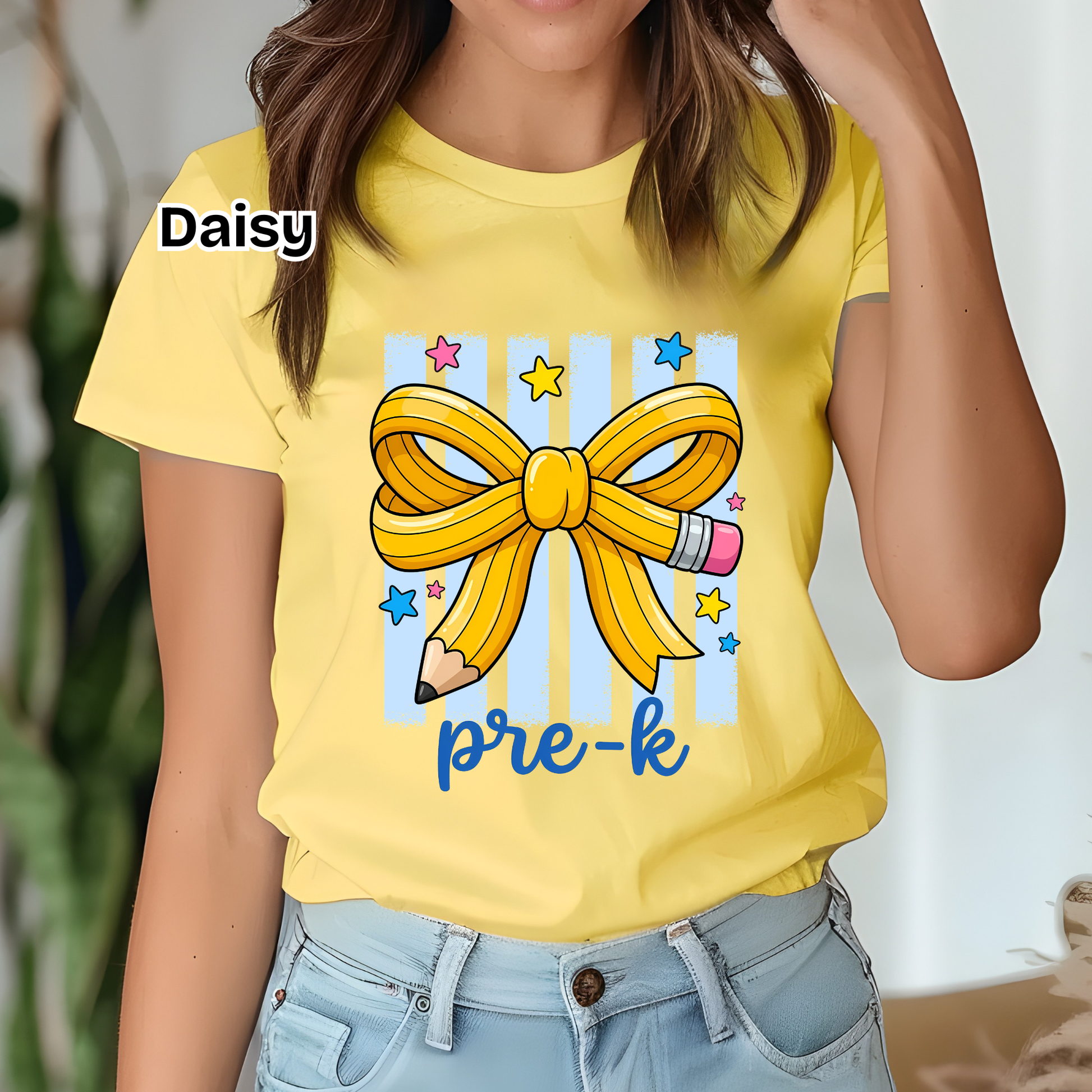 A woman wearing a bright yellow t-shirt with a graphic of a yellow pencil ribbon bow, blue and yellow stripes, stars, and the text 'Daisy pre-k'.