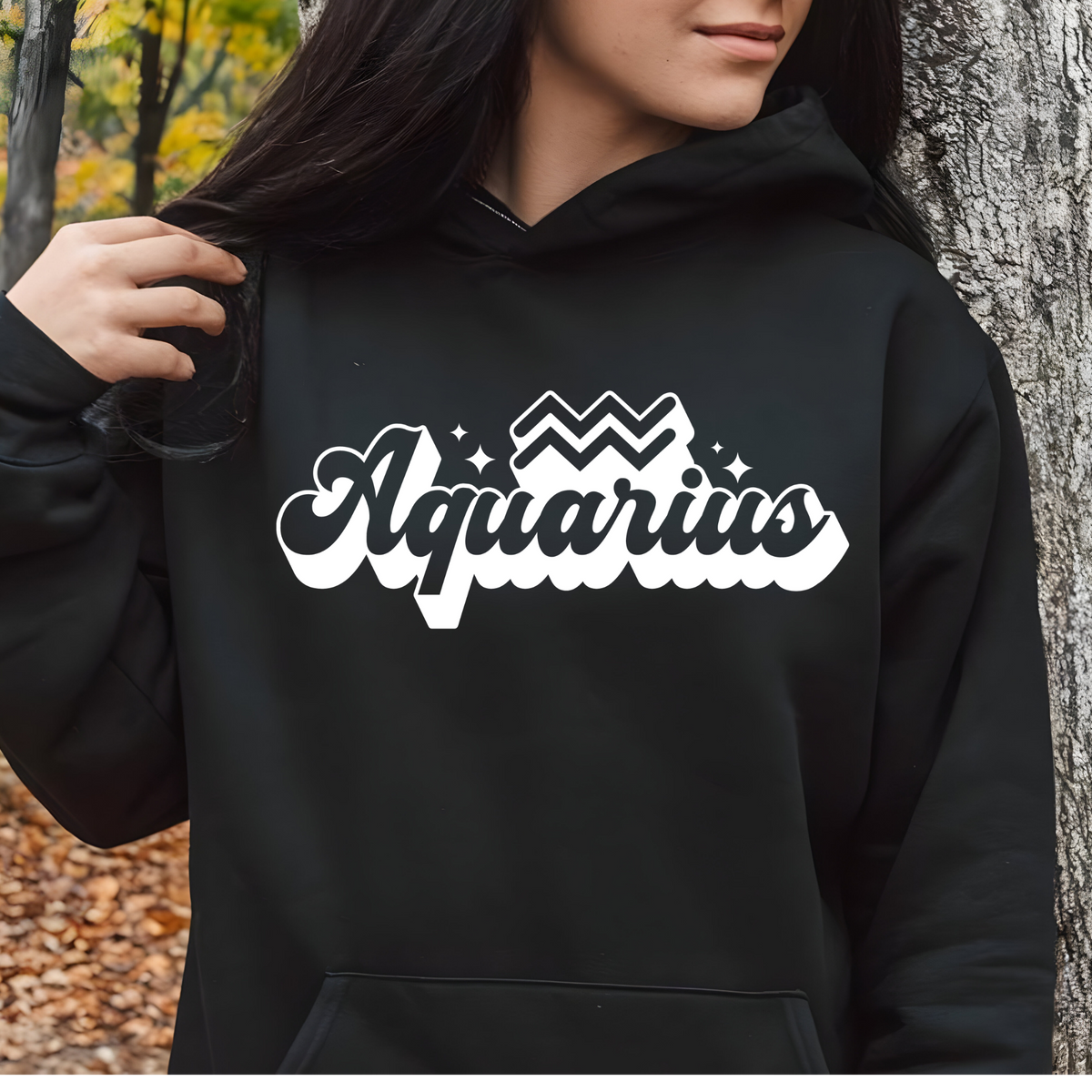 A person wearing a black hoodie with a large white 'Aquarius' graphic across the chest, photographed outdoors against a tree trunk and autumn ground.