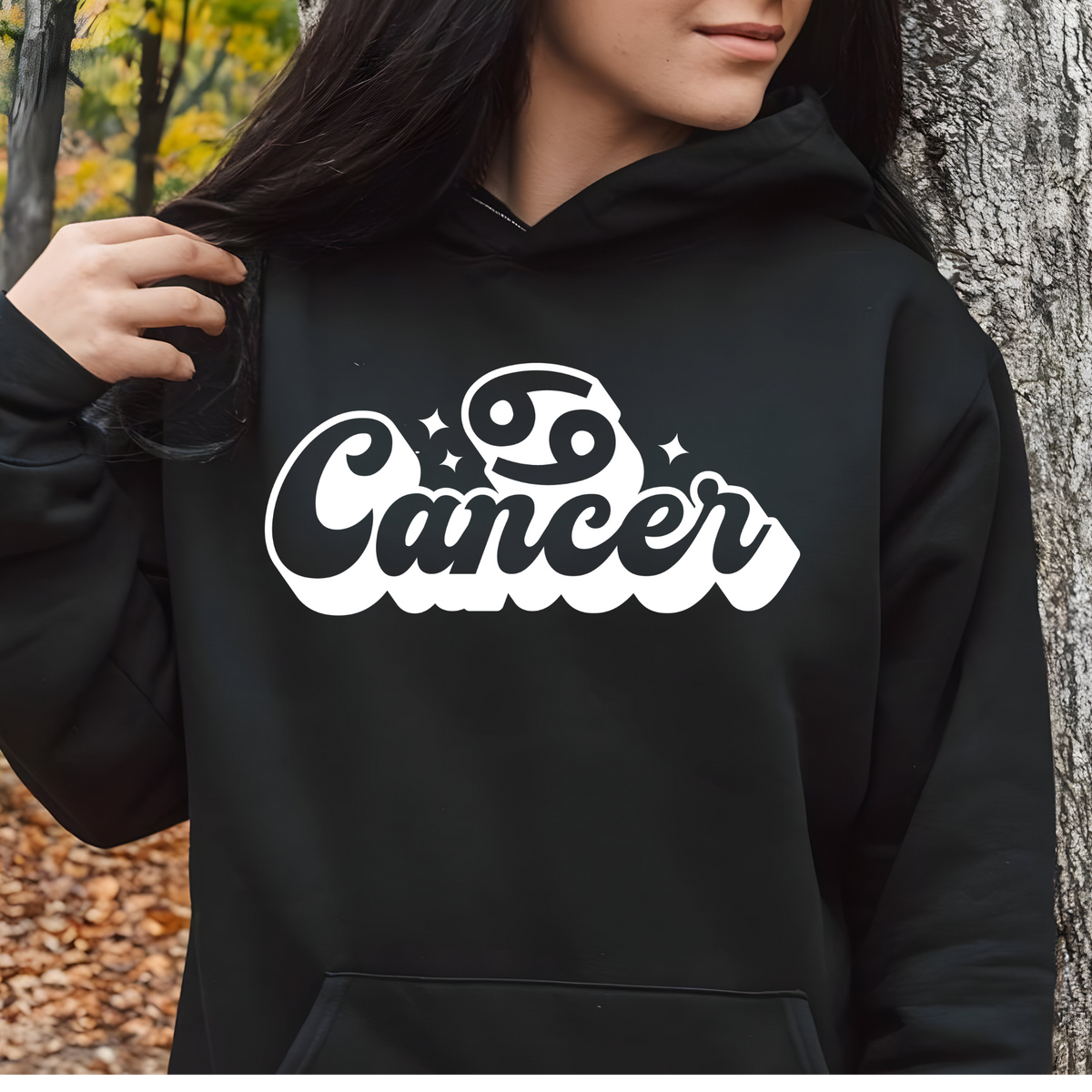 A person wearing a black pullover hoodie with a large white 'Cancer' zodiac sign graphic on the chest, photographed outdoors against a tree trunk and leaf-covered ground.