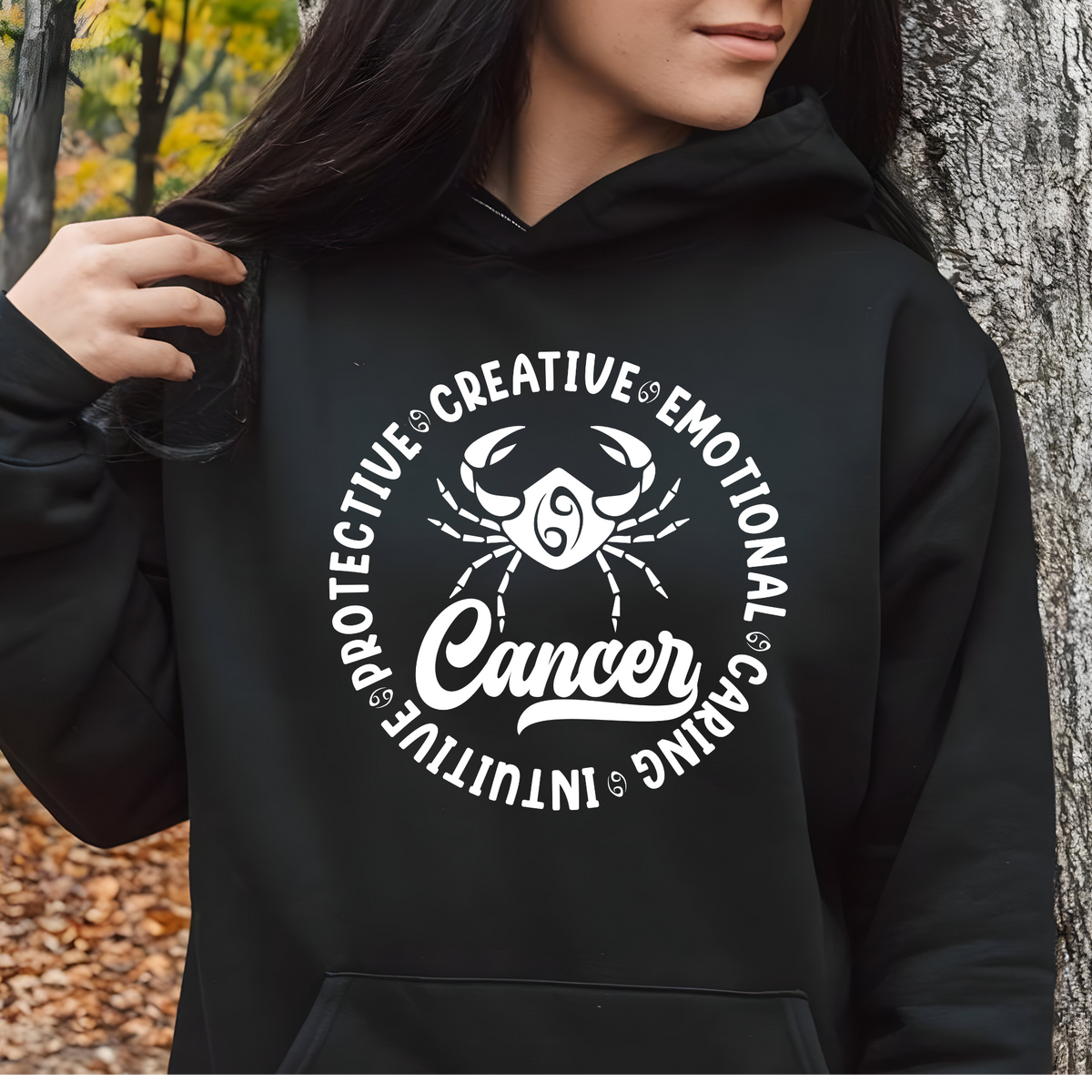 A person wearing a black hoodie with a large white Cancer zodiac graphic on the chest, standing outdoors next to a tree trunk.