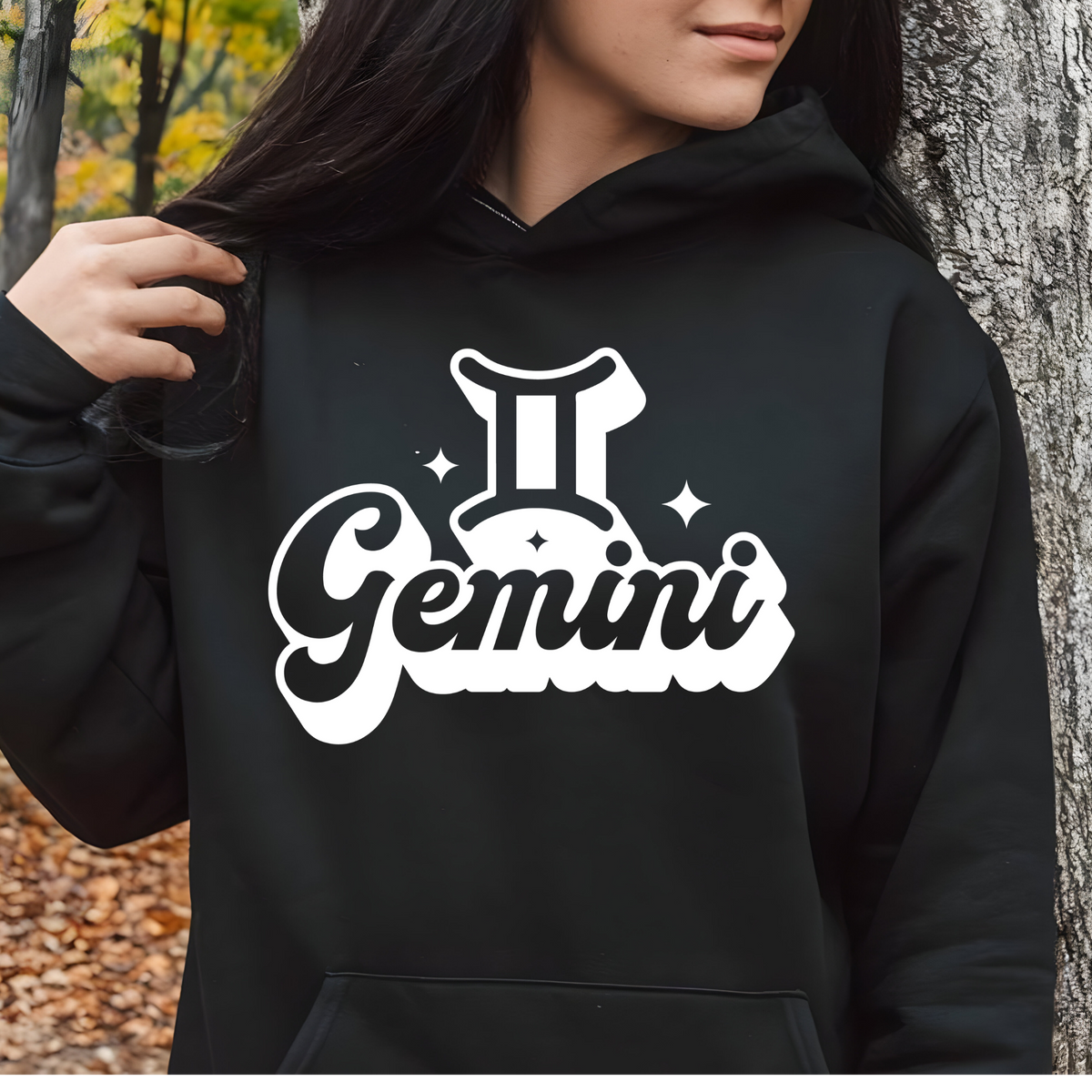 A person wearing a black hoodie with a large white Gemini zodiac graphic on the front, standing outdoors next to a tree trunk.