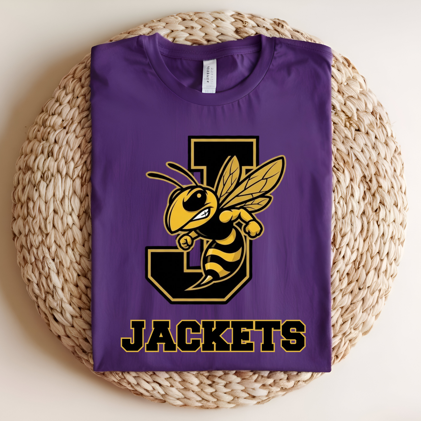 A folded purple t-shirt with a yellow and black bee mascot graphic over a large letter 'J' and the word 'JACKETS' printed below, displayed on a round woven straw mat.