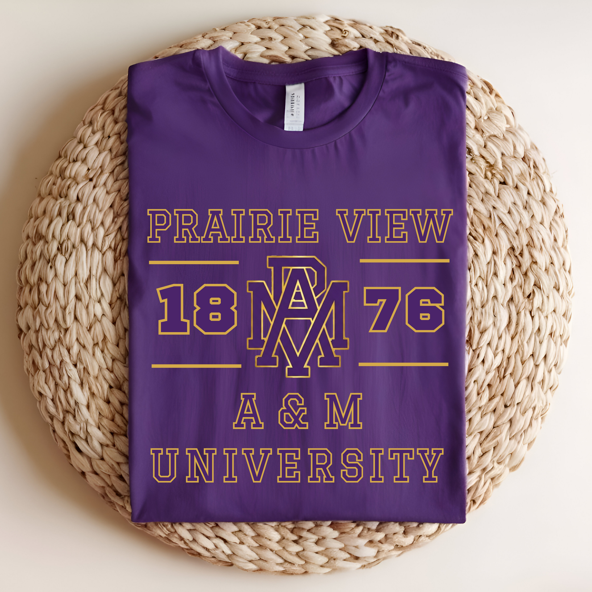 A folded purple t-shirt with gold and white text reading 'PRAIRIE VIEW 1876 A & M UNIVERSITY' and a large 'AM' monogram, displayed on a round woven straw mat against a light beige background.
