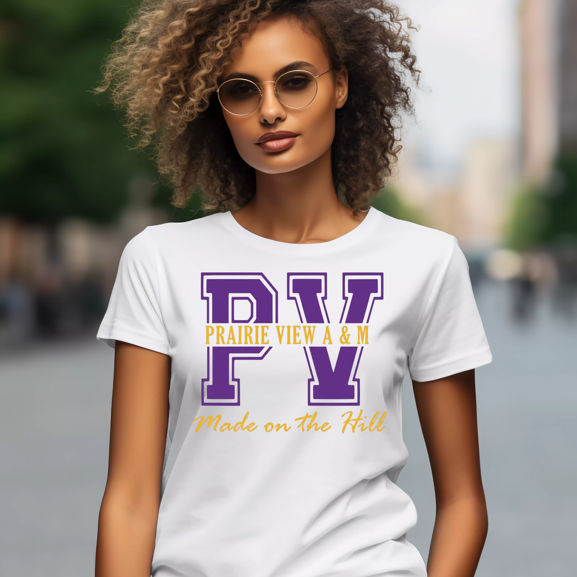 A woman wearing round sunglasses and a white graphic T-shirt with large purple 'PV' letters, yellow 'PRAIRIE VIEW A & M' text, and yellow 'Made on the Hill' script, photographed outdoors with a blurred city background.