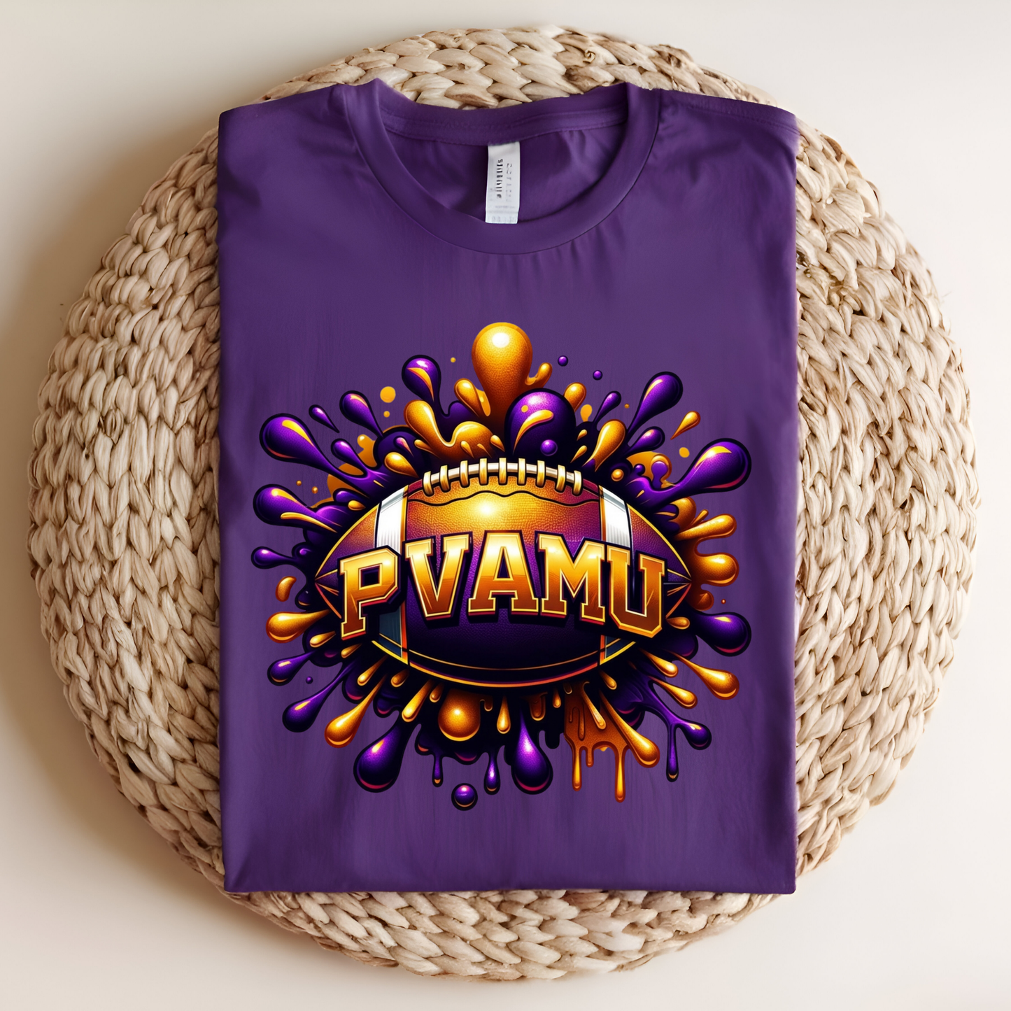 A folded purple t-shirt with a graphic design featuring a football and the text 'PVAMU' in gold and purple paint splatter, displayed on a round woven straw mat.