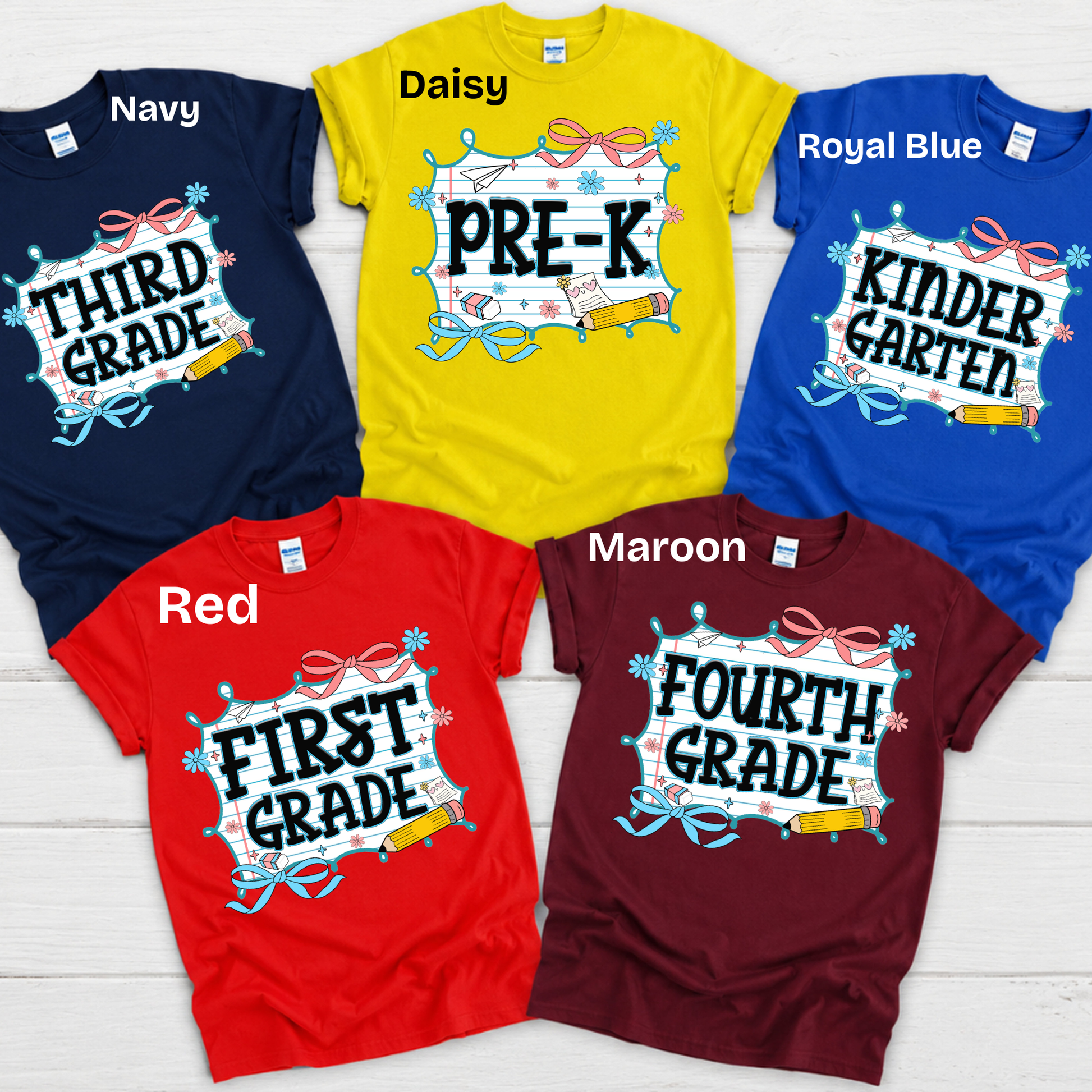 Five children's t-shirts in different colors are arranged in a flat-lay on a white wood background. The shirts are navy, yellow (labeled Daisy), royal blue, red, and maroon (labeled Maroon), each with a custom graphic design celebrating a milestone grade or 'First Grade' birthday.