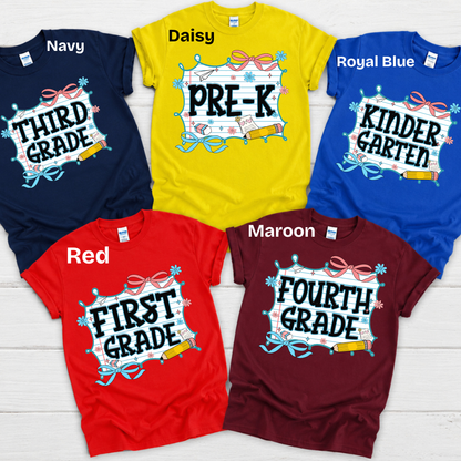 Five children's t-shirts in different colors are arranged in a flat-lay on a white wood background. The shirts are navy, yellow (labeled Daisy), royal blue, red, and maroon (labeled Maroon), each with a custom graphic design celebrating a milestone grade or 'First Grade' birthday.