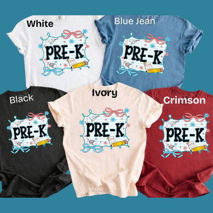 Five short-sleeve T-shirts in different colors are arranged on a teal background, each labeled with a color name: White, Blue Jean, Black, Ivory, and Crimson. Each shirt features a large front graphic with the text 'PRE-K' in black outlined letters inside a decorative white banner with pink bows, blue ribbon, and school-themed illustrations.