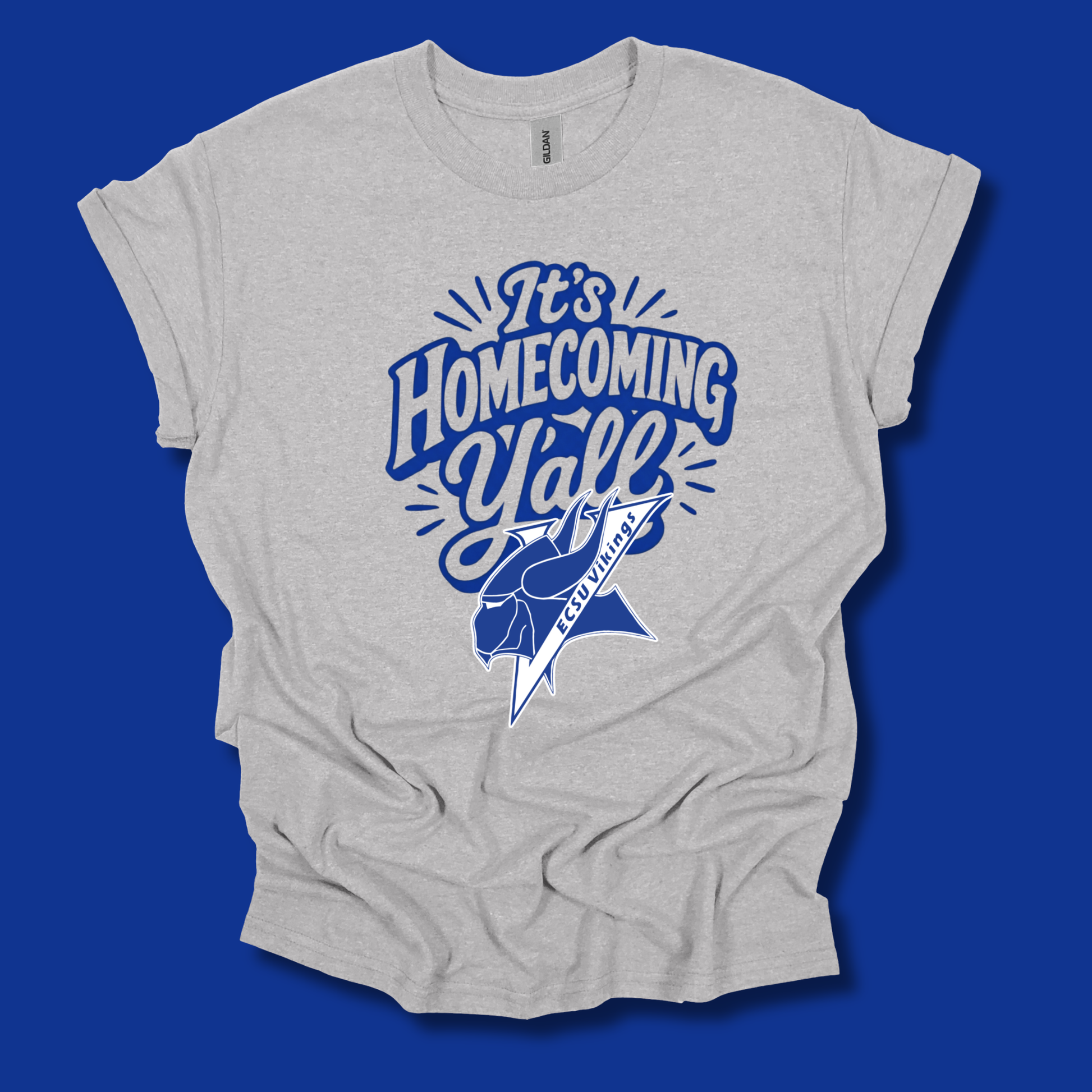 A light heather gray t-shirt with a blue graphic that reads 'It's Homecoming Y'all' and features a blue and white bobcat mascot, displayed against a solid blue background.