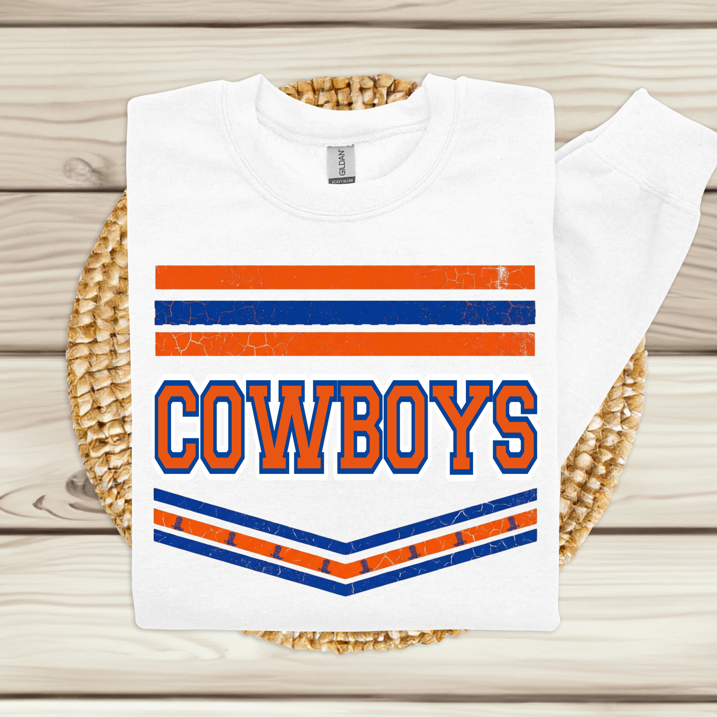 A white long-sleeve t-shirt with a distressed orange and blue graphic reading 'COWBOYS' on the front, displayed flat on a woven straw mat over a light wood plank background.