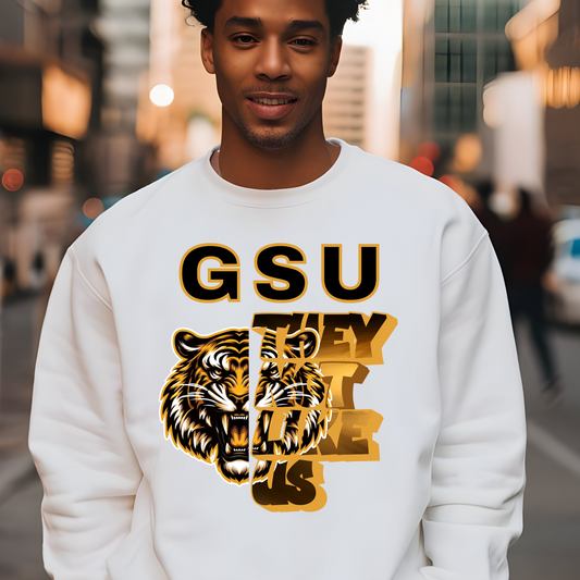 Person wearing a white sweatshirt with a tiger graphic and 'GSU THEY LIKE US' text on a city street background. Grambling Football:  GSU They Not Like Us Game Day Unisex Crewneck Sweatshirt