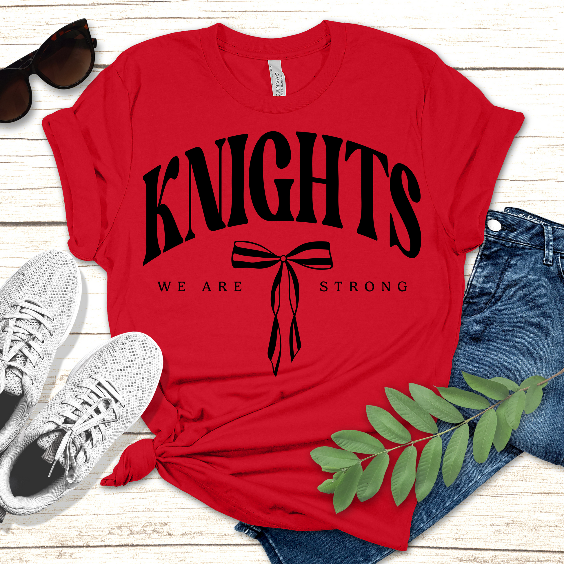 Red t-shirt with 'KNIGHTS WE ARE STRONG' text and graphics, paired with sunglasses, sneakers, and jeans on a wooden surface. Knights School Spirit Shirt - Knights We  Are Strong - Knights Spirit Tee