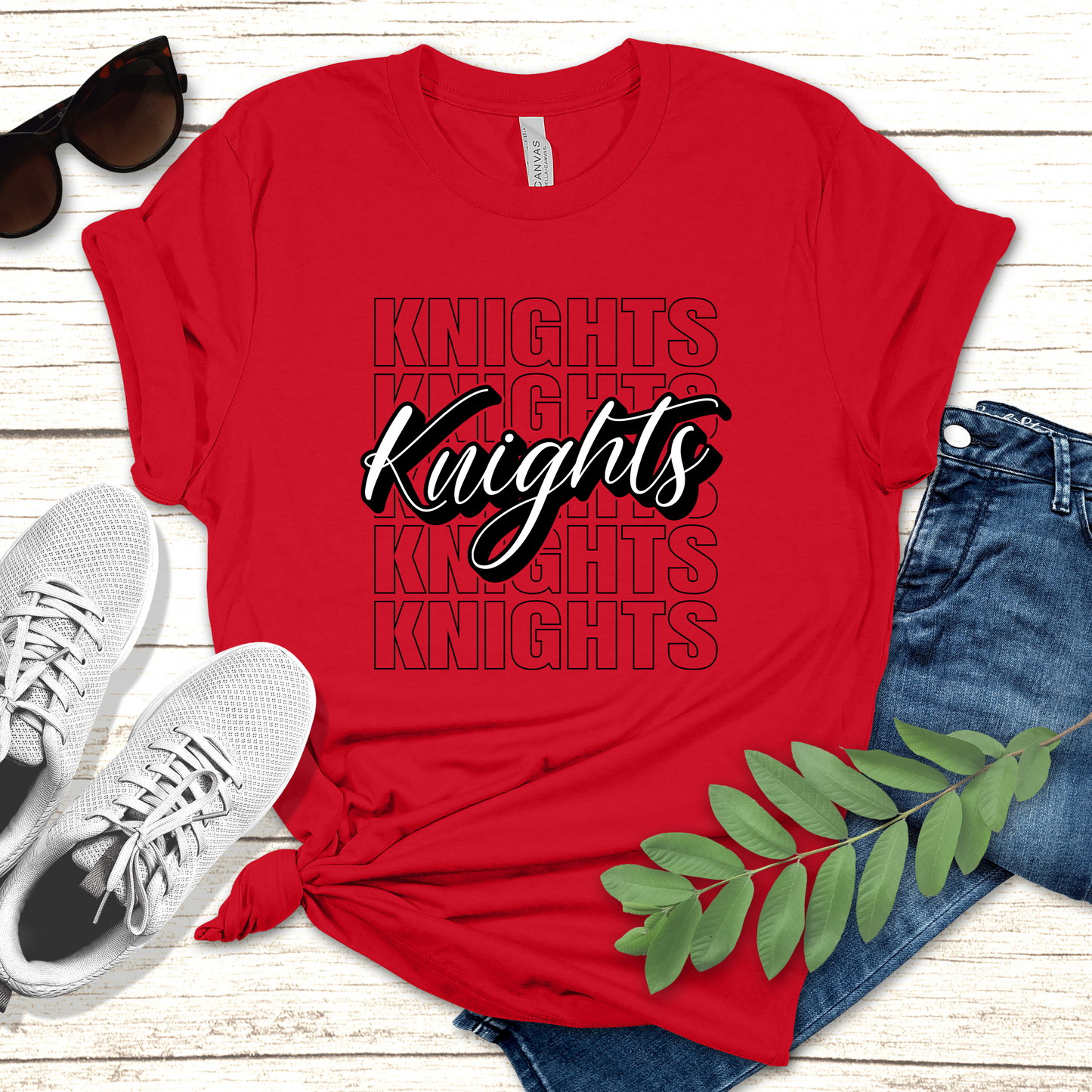 Red t-shirt with 'Knights' text, sunglasses, white sneakers, and jeans on a wooden surface. Knights School Spirit Shirt - Knights Cursive Stacked - School Pride T-Shirt