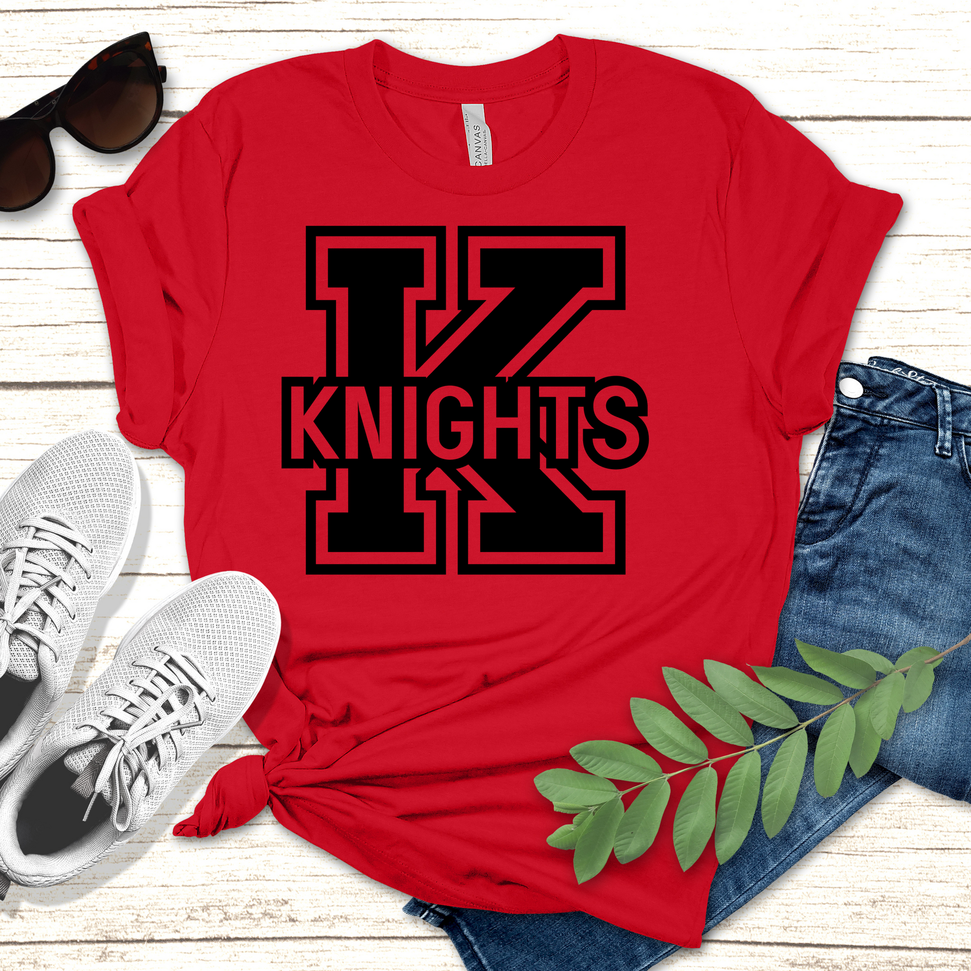 Red t-shirt with 'Knights' design, sunglasses, white sneakers, and jeans on a wooden surface. Knights School Spirit Shirt - Knights Logo: Loud & Proud Knights