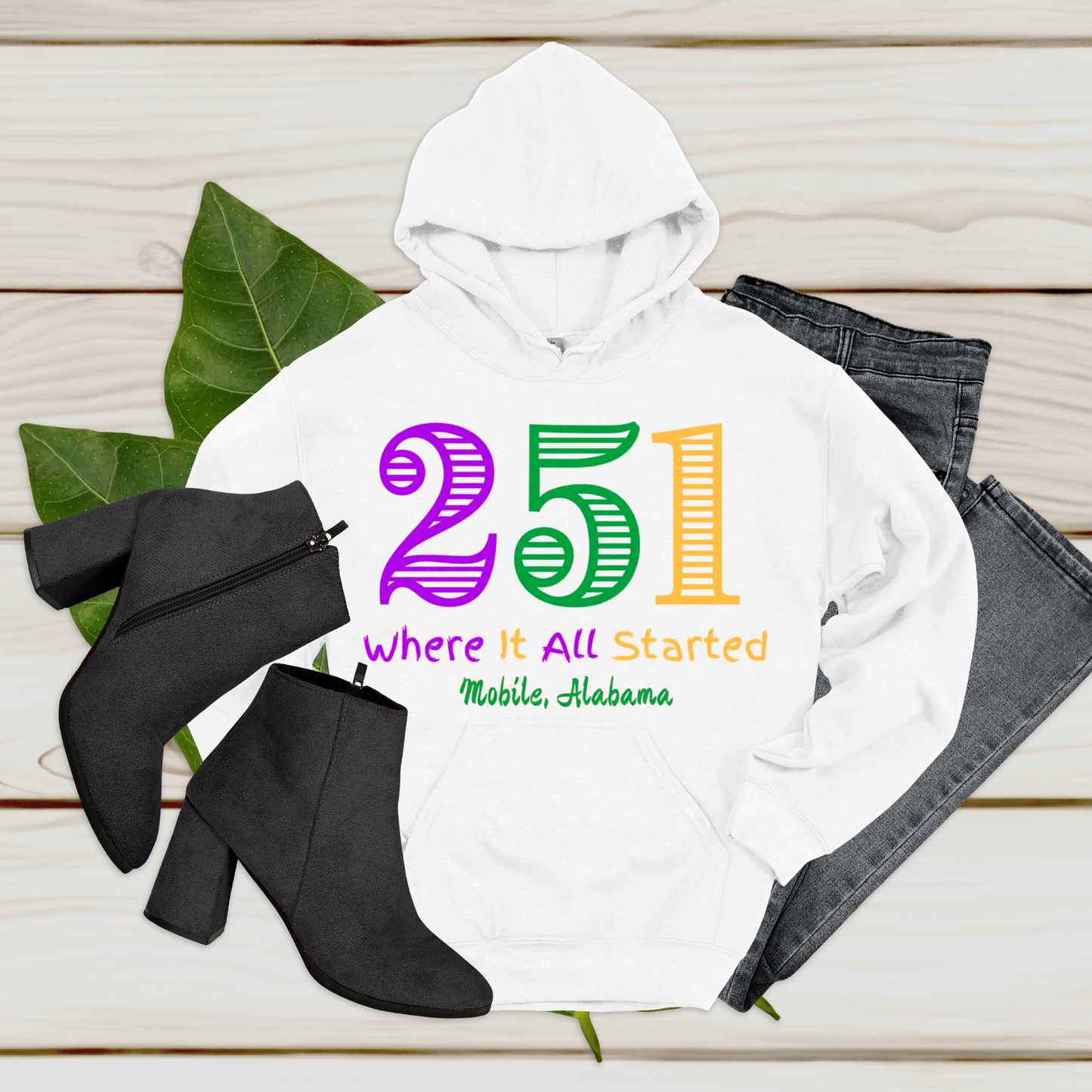 White hoodie with colorful text '251' and 'Where It All Started Mobile, Alabama' on a wooden surface with black boots and jeans.