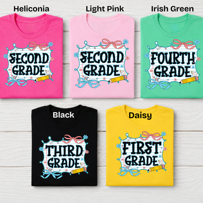 Five folded graphic T-shirts arranged in a 2x3 grid on a light wood background, labeled with color names at the top: Heliconia pink, Light Pink, Irish Green, Black, and Daisy yellow. Each shirt has a school-grade-themed graphic with text such as SECOND GRADE, FOURTH GRADE, THIRD GRADE, and FIRST GRADE.