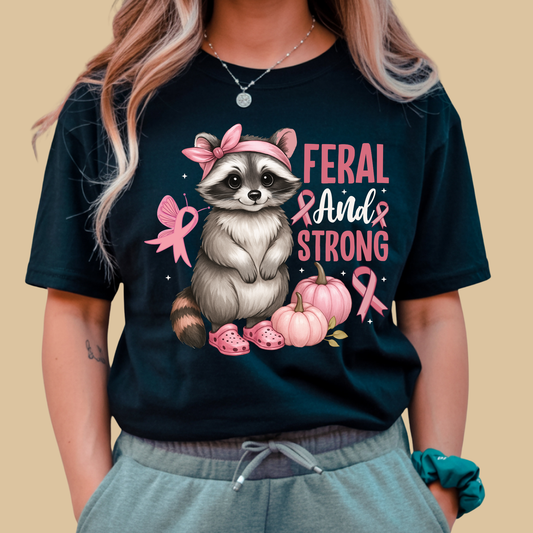 Person wearing a black t-shirt with a raccoon graphic and 'Feral and Strong' text on a beige background Pink Ribbon Tee: Feral and Strong - Supportive Awareness T-shirt