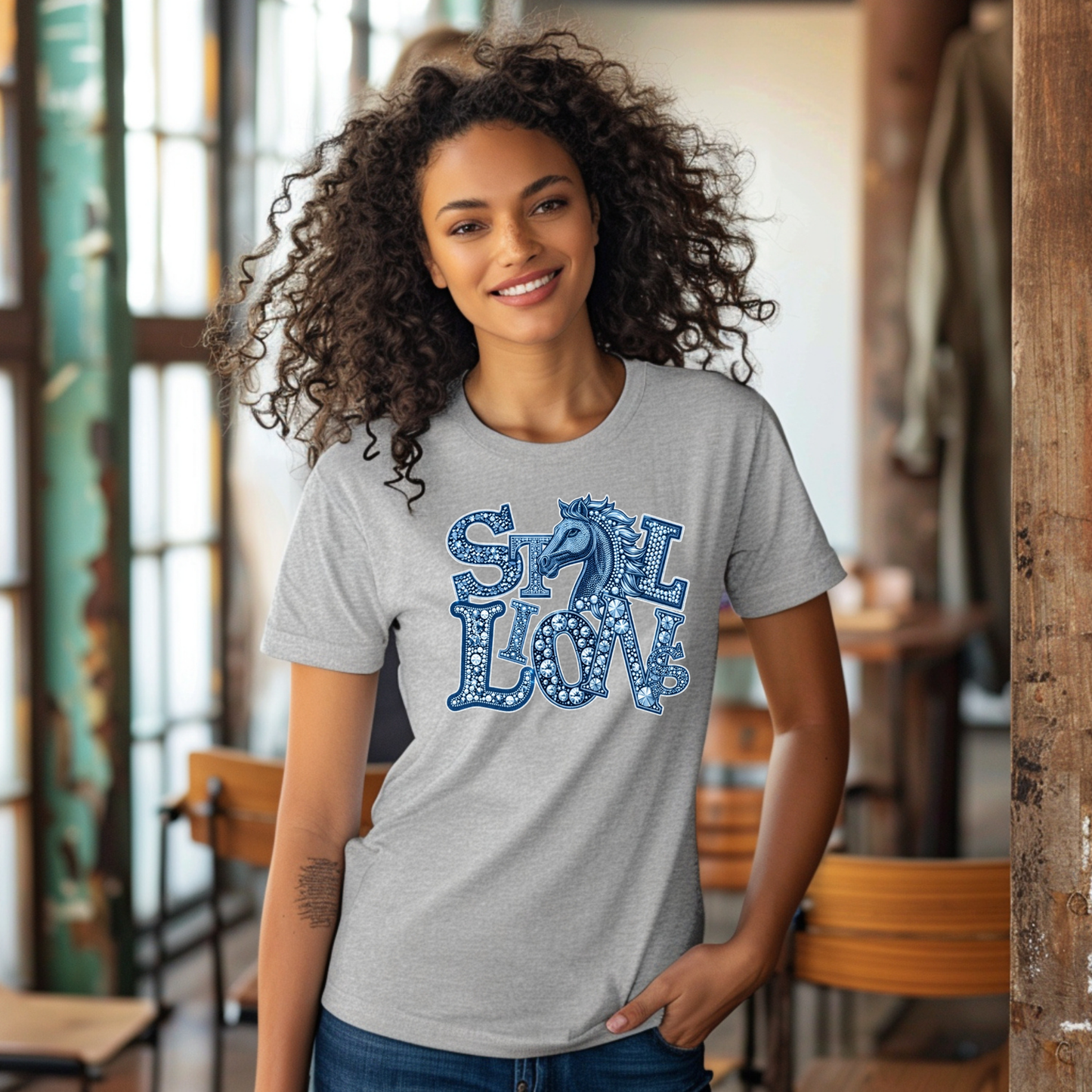 A woman with curly dark hair wearing a heather gray t-shirt with a blue and white 'STALLIONS' graphic and horse logo, standing in a rustic indoor cafe setting.