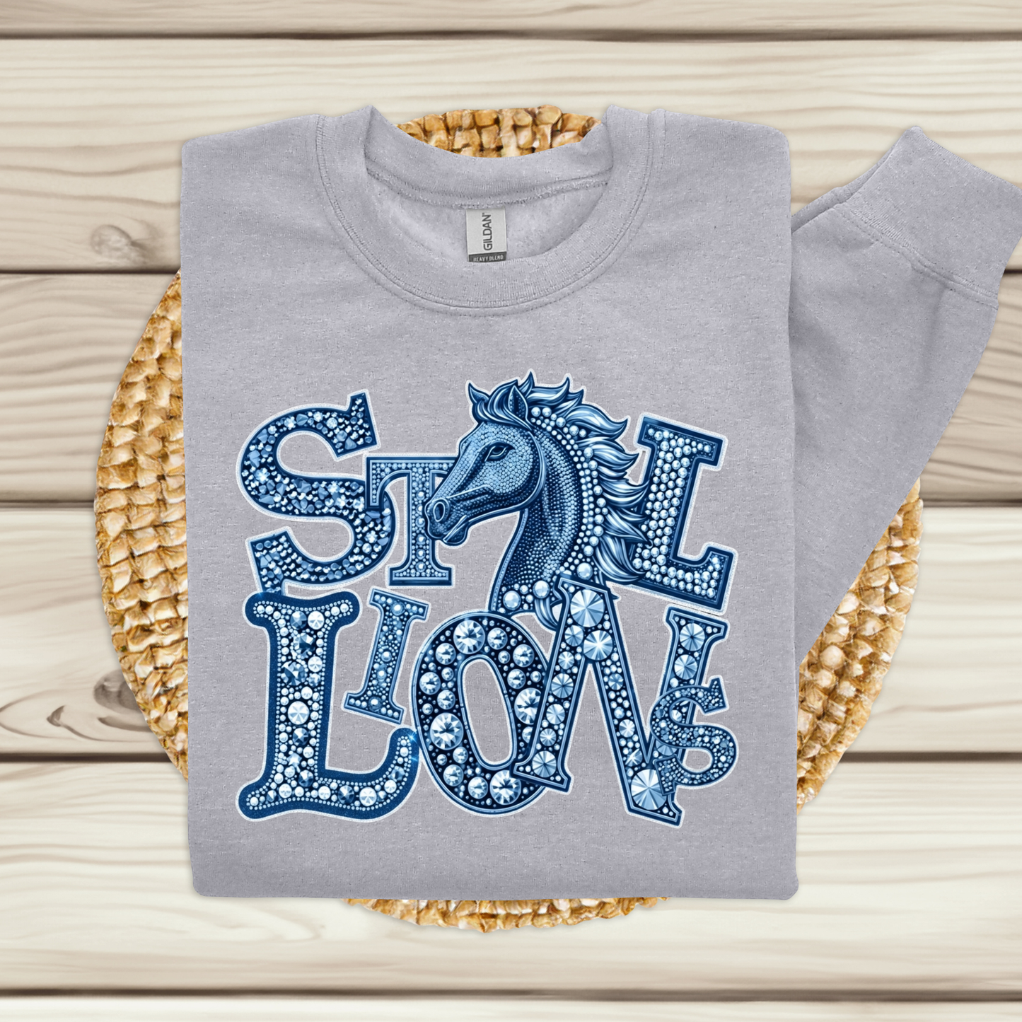 A light gray sweatshirt laid flat on a woven straw mat over a light wood plank surface. The sweatshirt has a large blue graphic reading 'STILL LOVE' with a horse head illustration integrated into the lettering.