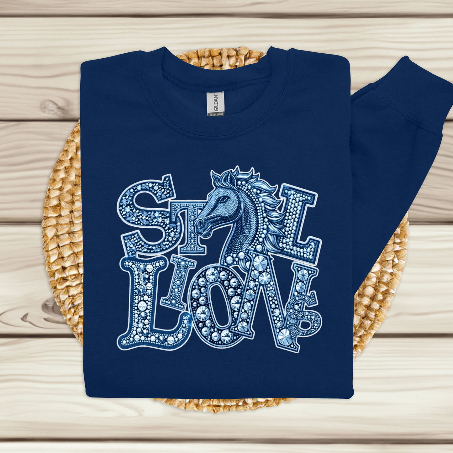 A navy blue sweatshirt with a decorative graphic reading 'STILL LOVE' in white and light blue, featuring a horse head motif, laid flat on a woven straw mat over a light wood plank surface.
