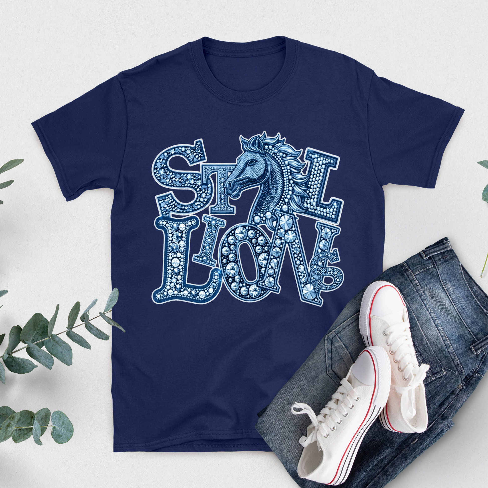 A navy blue short-sleeve graphic T-shirt laid flat on a light background, styled with blue jeans, white sneakers, and green leaves. The shirt features a large decorative graphic reading 'STILL LOVE' with a horse head motif in light blue and white.