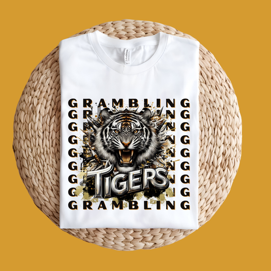 A folded white graphic T-shirt displayed on a round woven straw mat against a solid mustard-yellow background. The shirt features a large central tiger head illustration with the word 'TIGERS' in bold lettering, surrounded by repeated 'GRAMBLING' text in black.