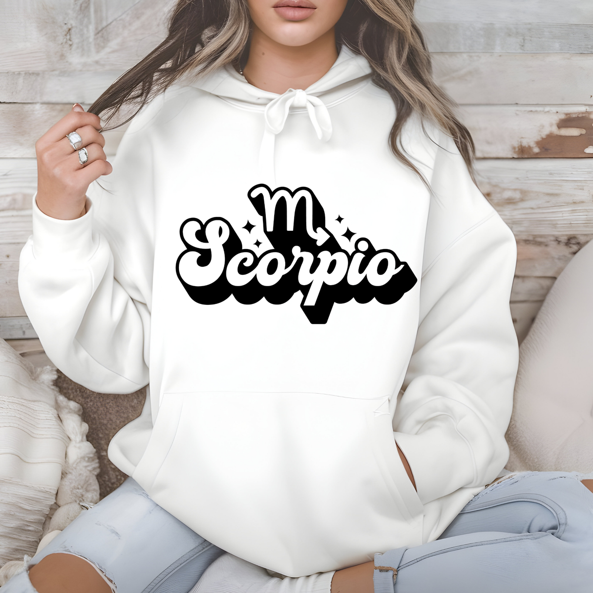 A woman wearing a white pullover hoodie with a black 'My Scorpio' graphic printed on the chest, sitting against a rustic wood panel background.