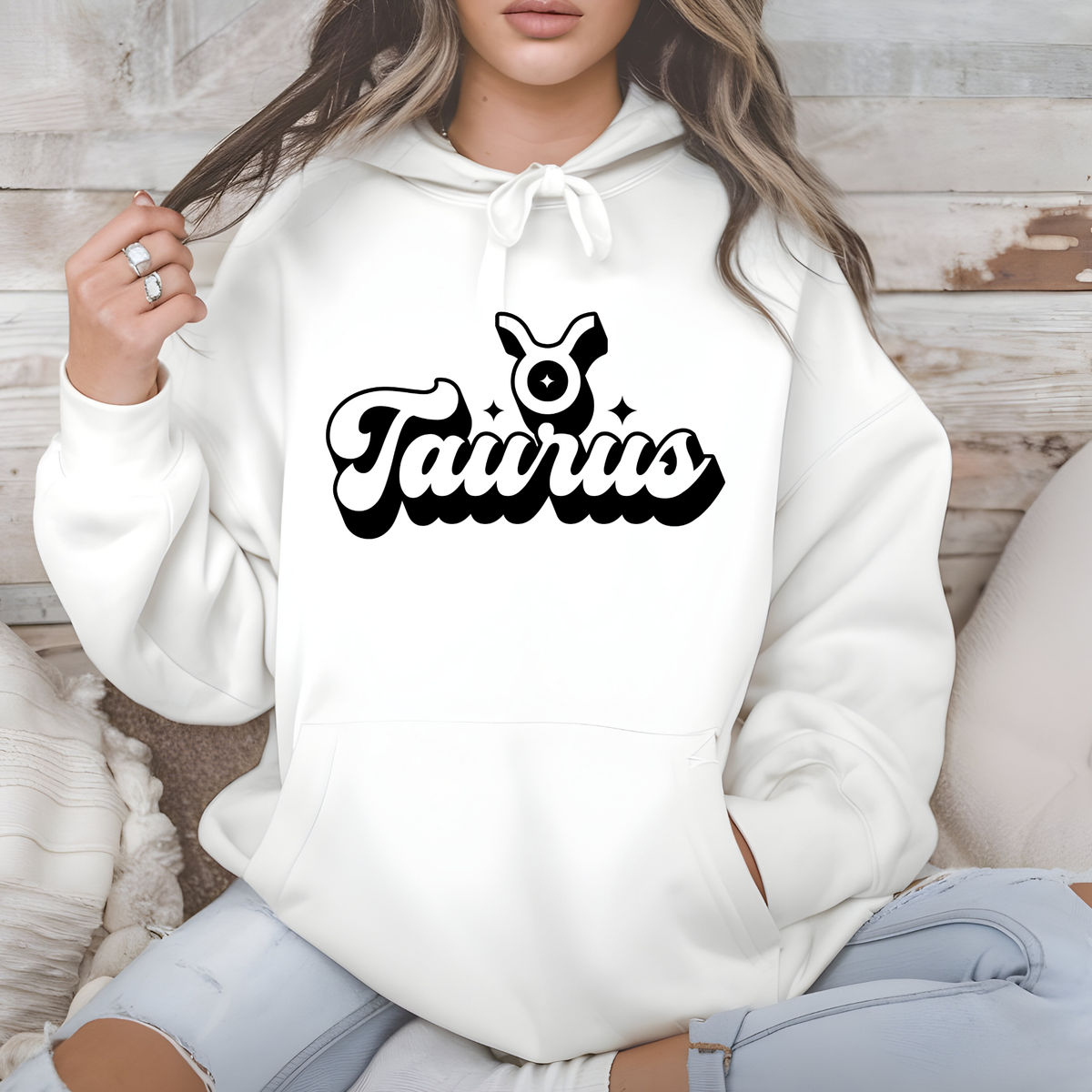 A woman wearing a white pullover hoodie with a black 'm Virgo' graphic on the chest, sitting against a rustic wood and neutral background.