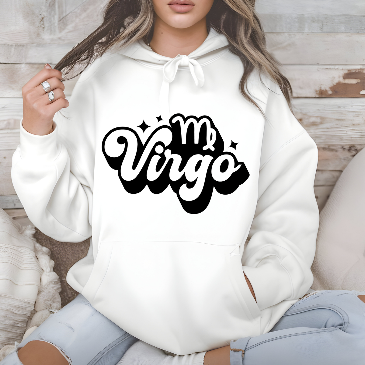 A woman wearing a white pullover hoodie with a black 'm Virgo' graphic on the chest, sitting against a rustic wood and neutral background.