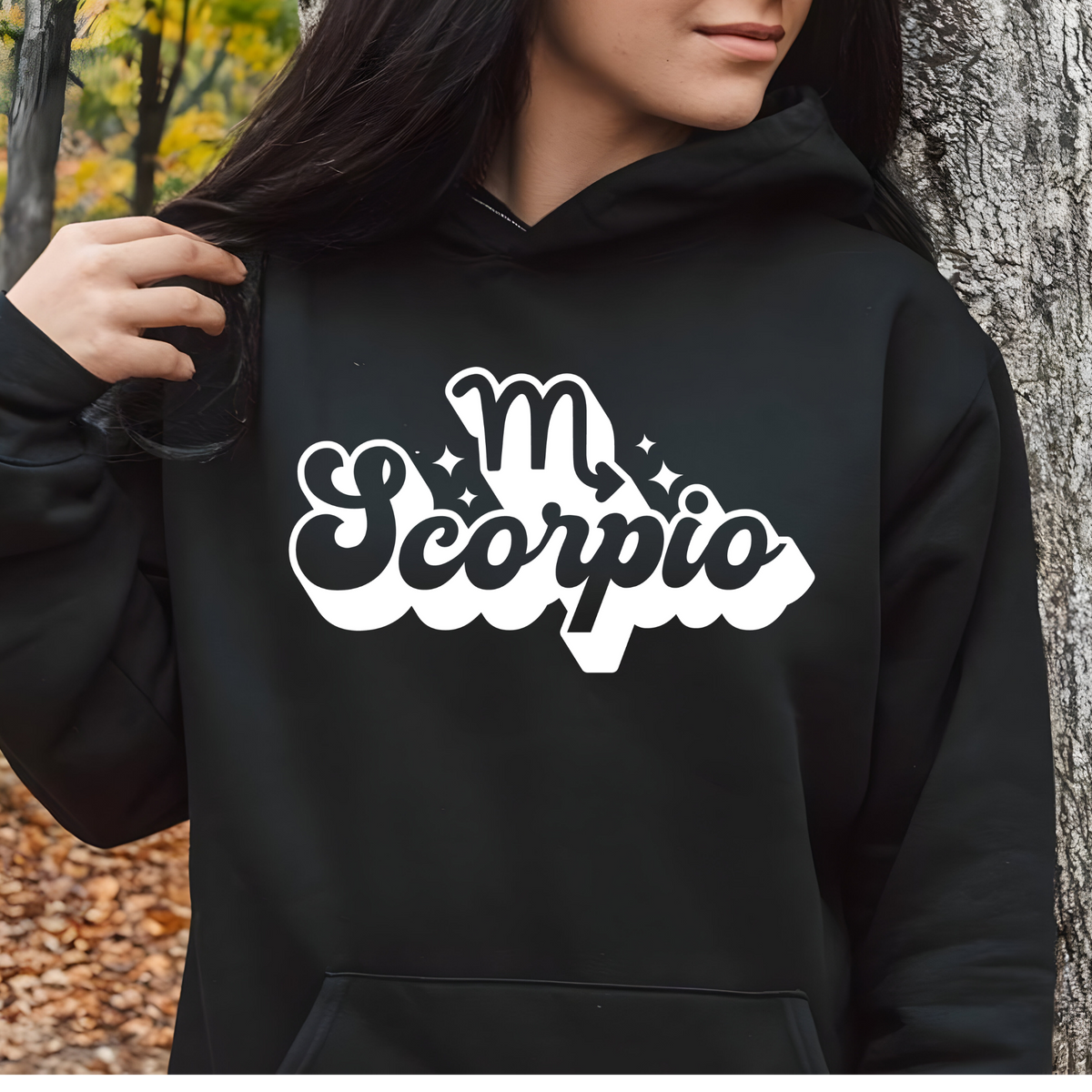 A person wearing a black hoodie with a white 'm Scorpio' graphic on the chest, standing outdoors next to a tree trunk.